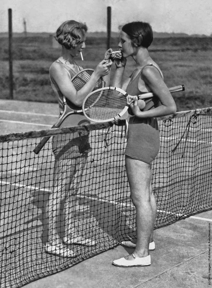 #19 Two women lighting cigarettes on a tennis court in Essex, England 1930s.