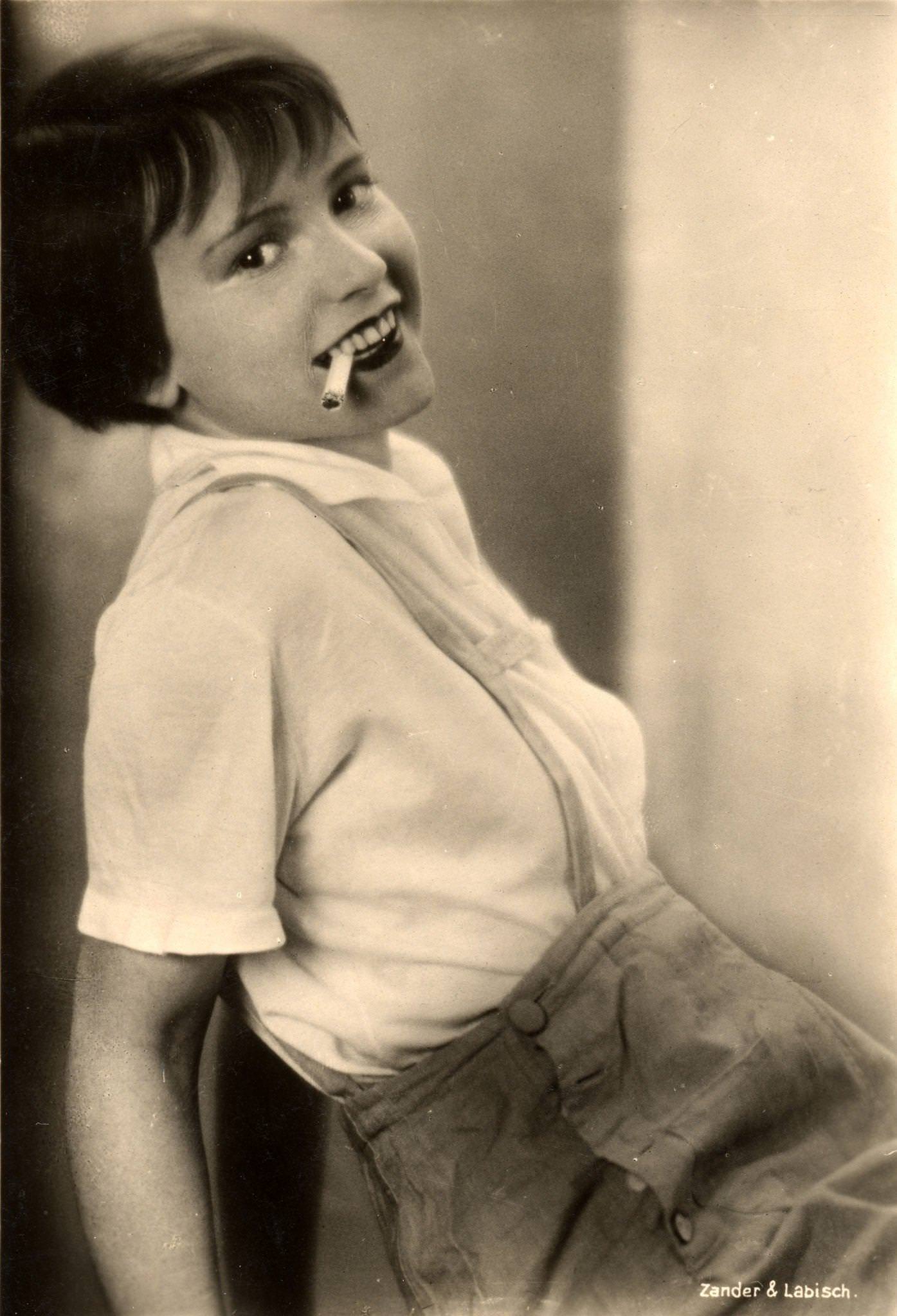 #20 American actress Louise Brooks poses in lederhosen and smokes a cigarette in this postcard image from Germany, 1920s or 1930s.