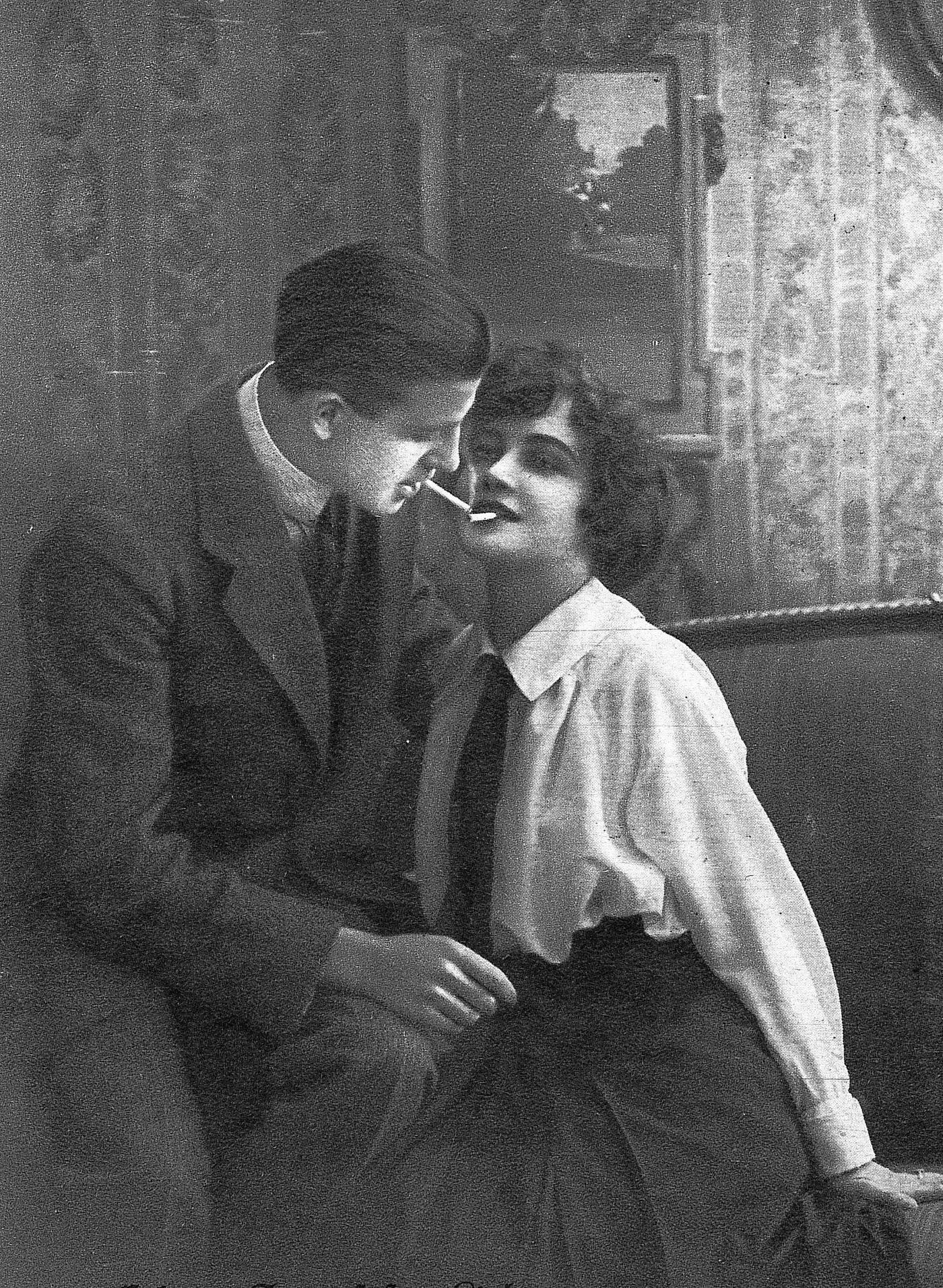 #32 Young man bends over to a woman sitting on the sofa, lights his cigarette with the glow of hers, 1930.