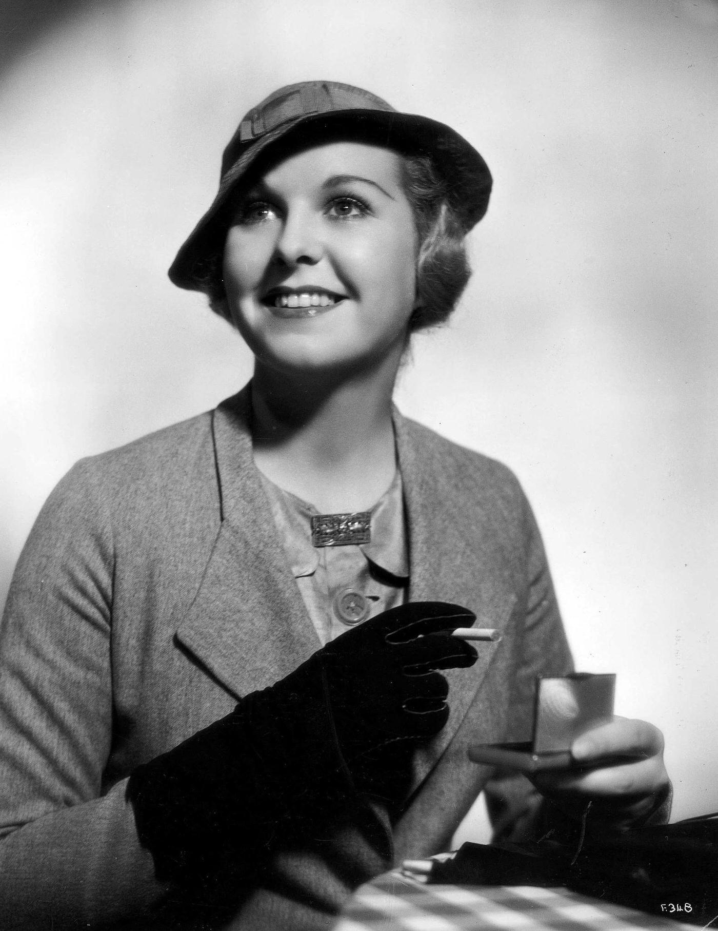 #62 A lady wearing a grey flannel suit with a stitched hat and black gloves about to light up a cigarette, 1935