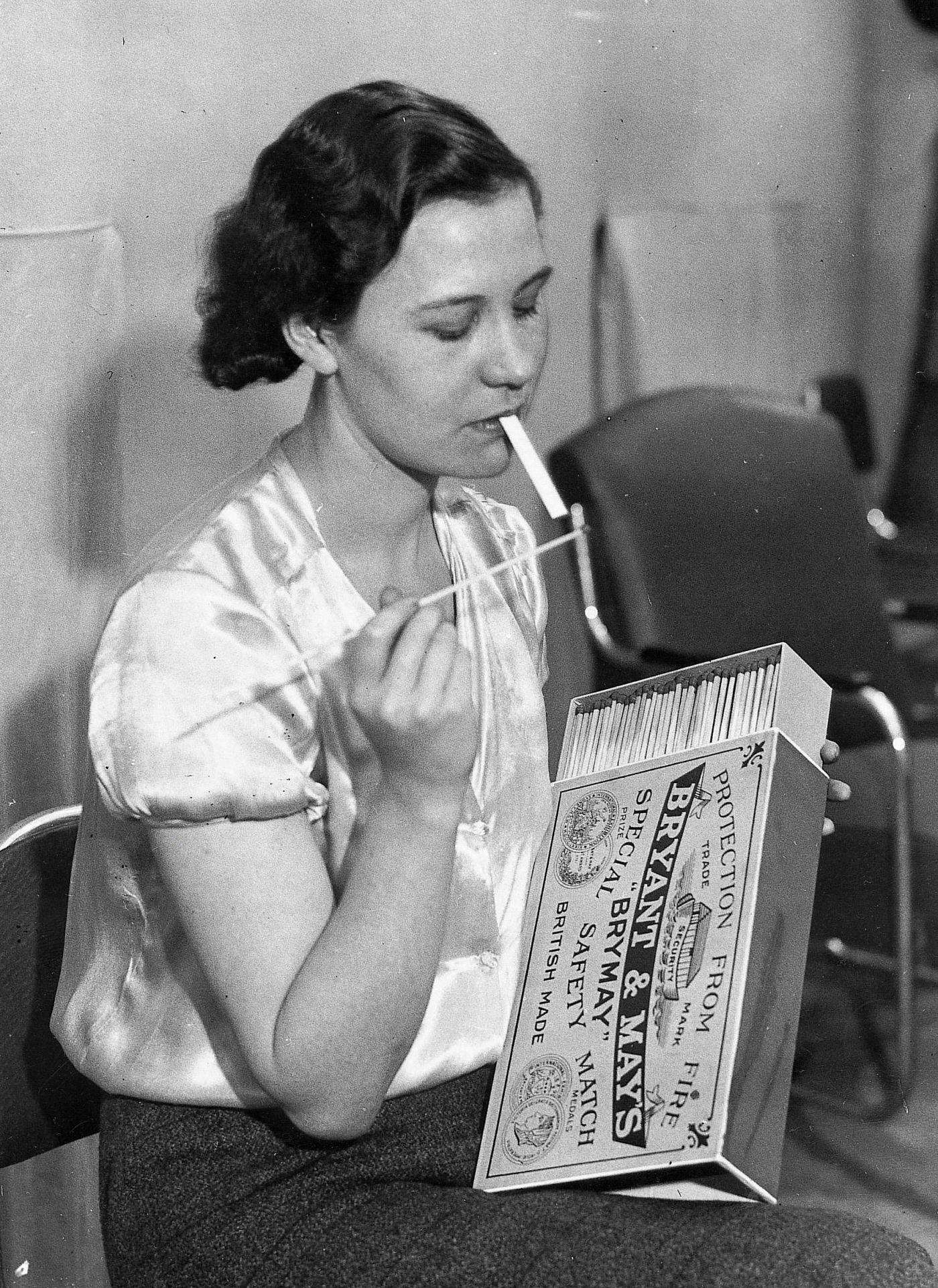 #63 A woman is lighting a cigarette with oversized matches at the 39th International Tabacco Exhibition, London, 1935