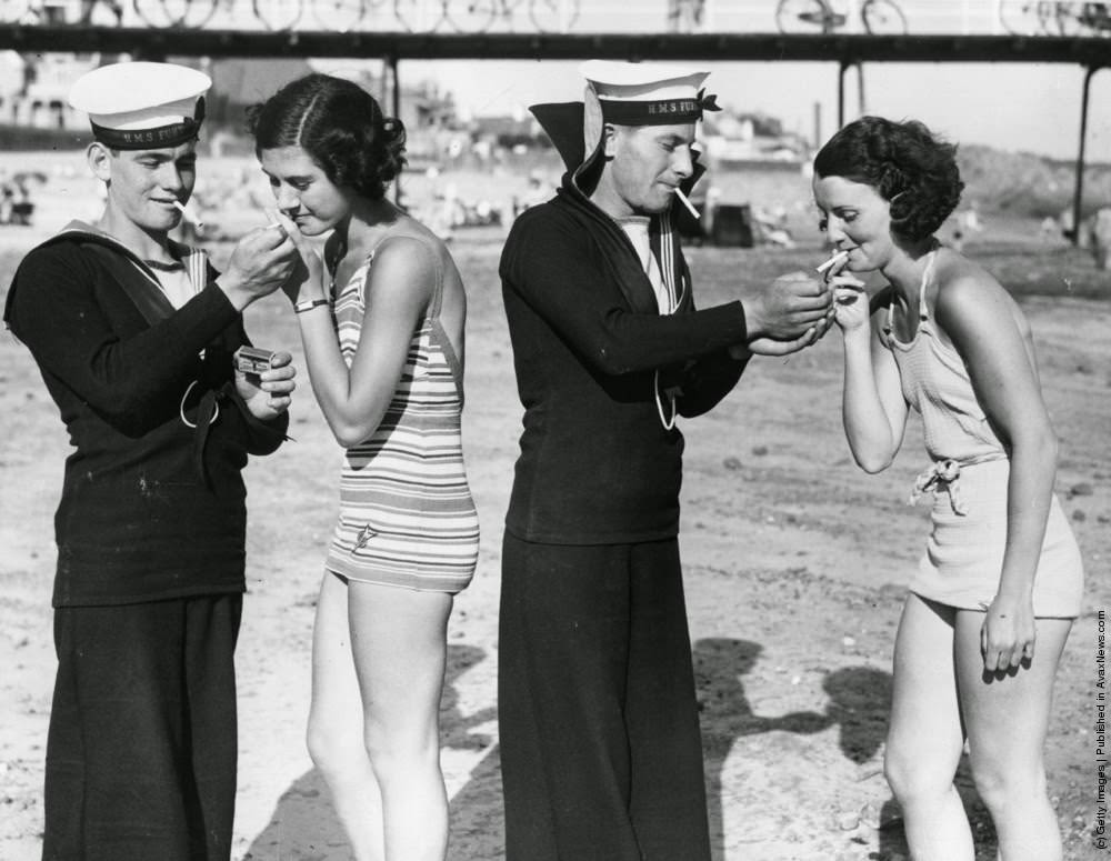 #14 Two sailors from HMS Fury enjoy a smoke with two women in bathing suits on a beach in Jersey, where their ship is on a visit, 1935