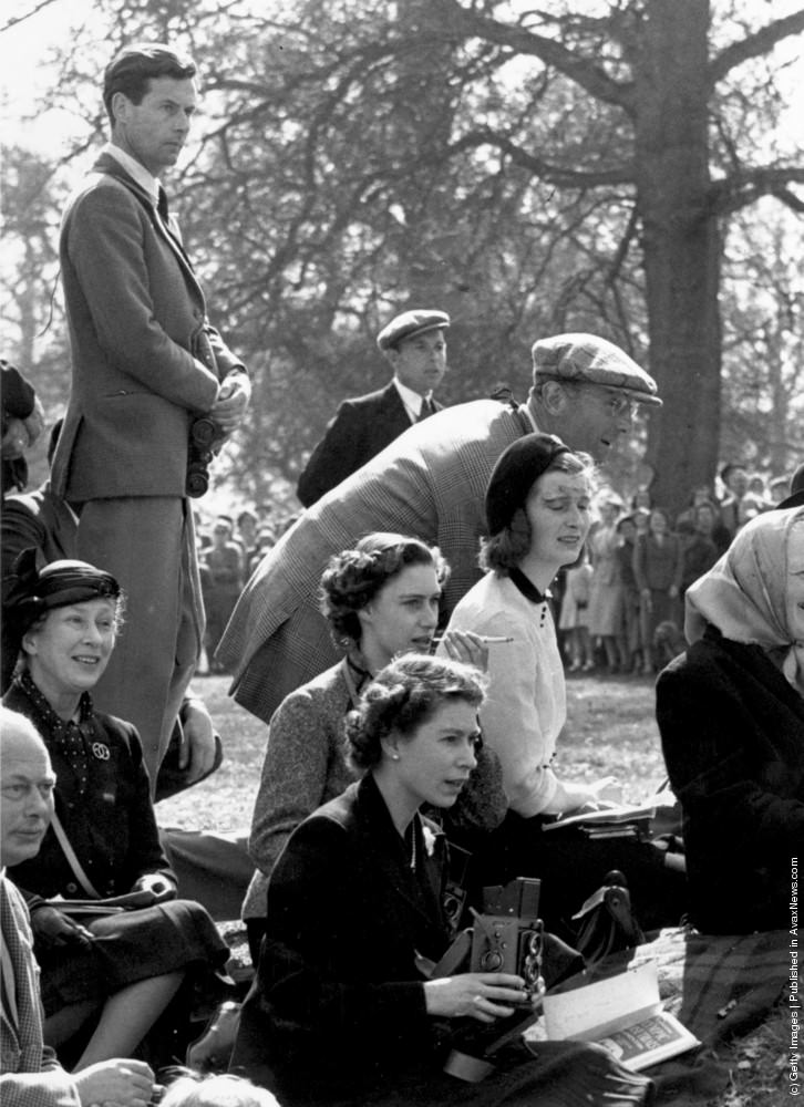 #15 Queen Elizabeth II holding a camera at the Olympic Horse Trials at Badminton, whilst Princess Margaret sits behind her, smoking a cigarette and watching the action. Also in the group is Group Captain Peter Townsend (left). 13th October 1955.