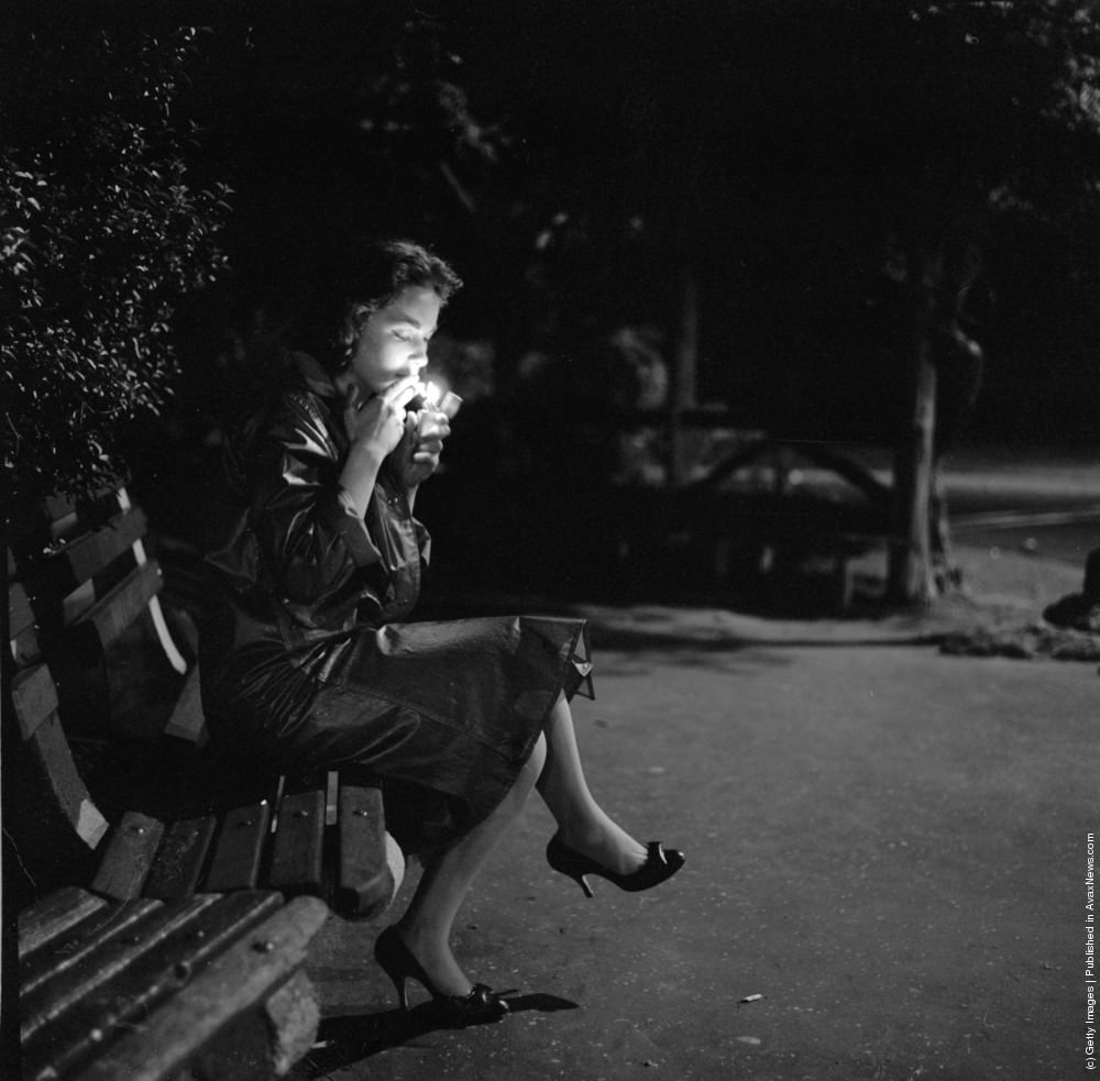 #20 A young woman lighting a cigarette as she sits on a New York park bench at night, circa 1957.