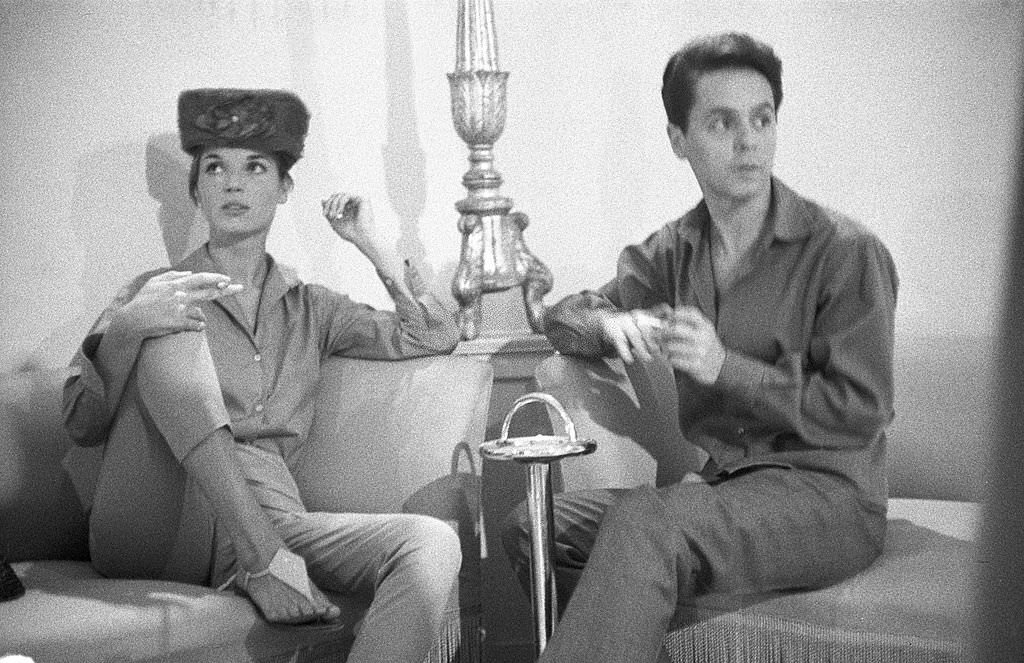 #25 Italian tailor Roberto Capucci sitting on a sofa with a model, both smoking cigarettes, Rome, 1950s.