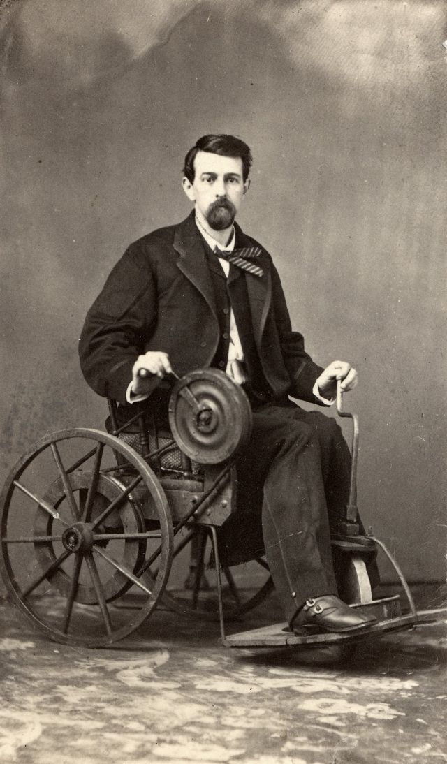 #9 A man sits in a wheel chair that features a flywheel with hand-crank that turns a belt and powers the large back wheels