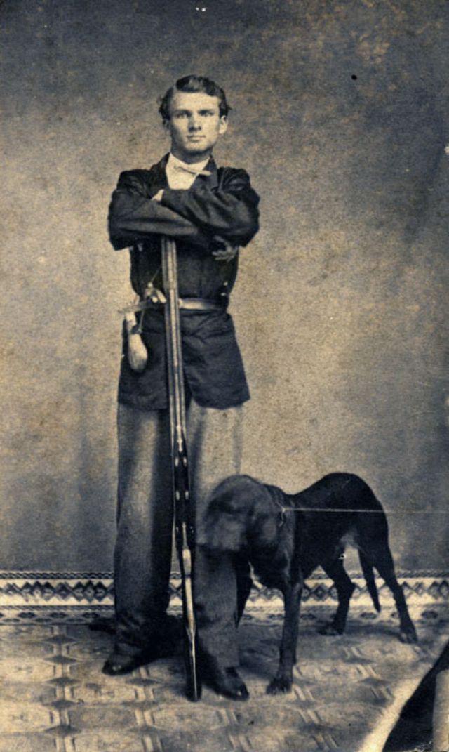 #14 A thin young man, likely in his late teens or early twenties, stands with forearms resting atop the muzzle of his firearm and his dog
