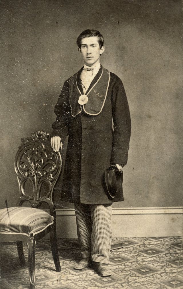 #16 A young man poses in his fraternal sash with his initials embroidered on one side and a flower in the center