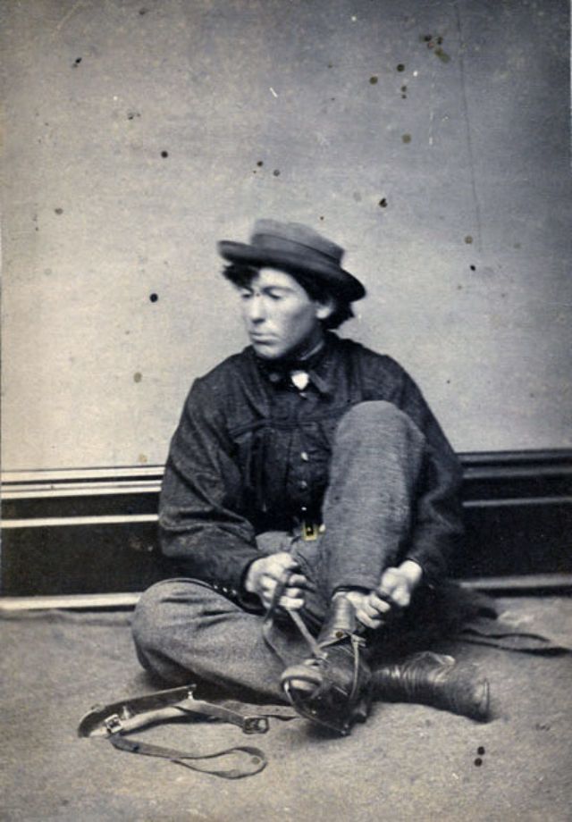 #17 A young man sits on a blanket upon the floor of the photographer’s studio, his left leg raised and hands pulling the leather straps that fasten an ice skate to his ankle-high boot