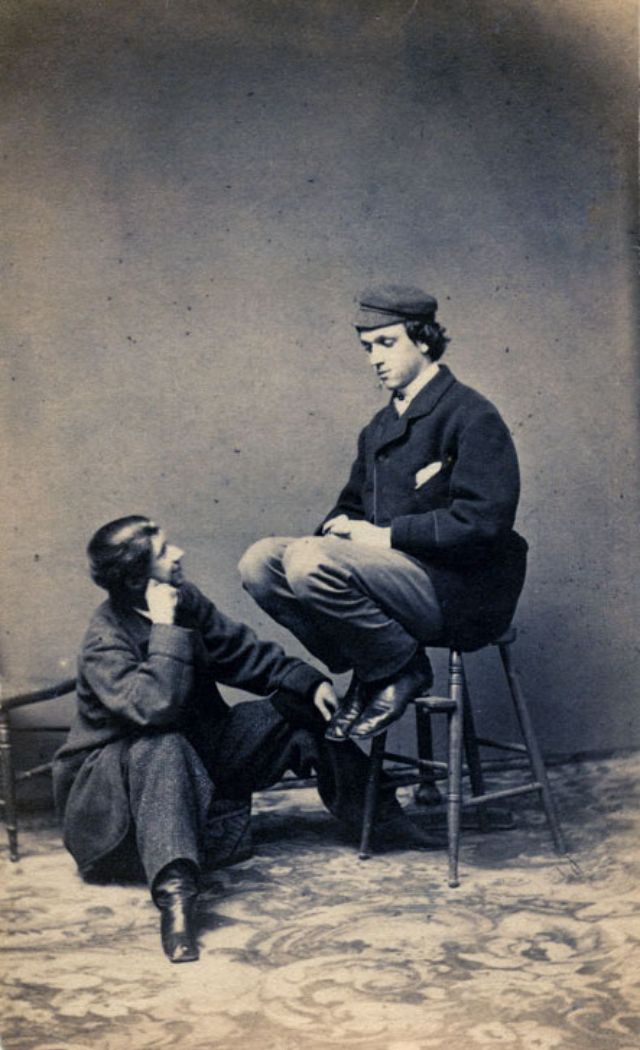 #18 A young man wearing a cap sits on a stool, looking down at another young man seated on a cushion and returning his gaze
