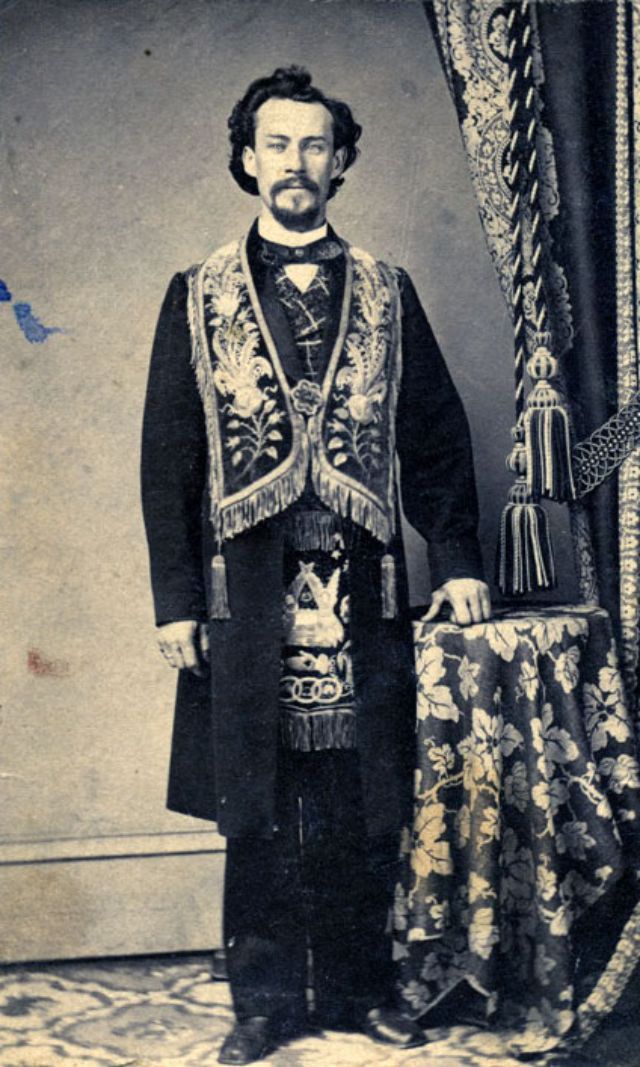 #1 A bearded man stands before the camera operator wearing the embroidered accouterments that signal his membership in the Masons