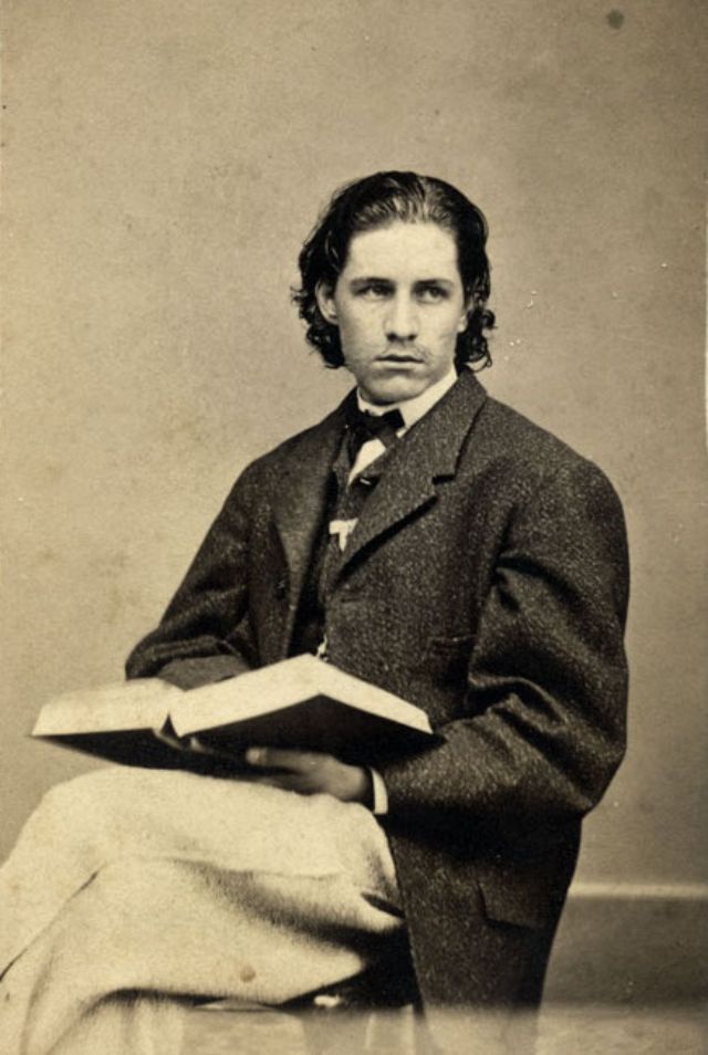 #19 A young man with a ribbon pinned to the lapel of his vest sits in a chair and holds a book