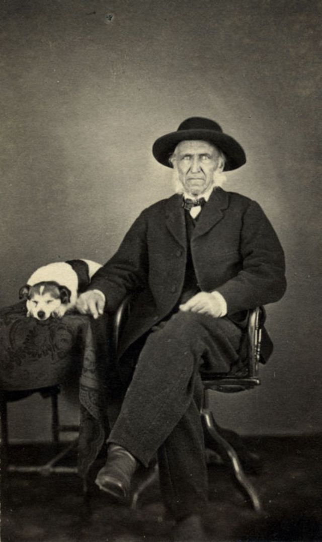 #20 An elderly man sits in a chair with his right arm resting on a cloth-covered table on which is seated a dog. The dog is asleep, head resting on paws