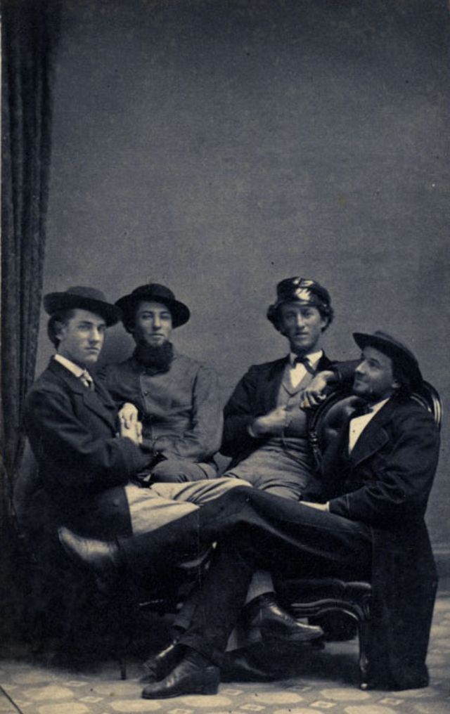 #27 Four young man, all with smiles or hints of smiles upon their faces, relax in chairs and a bench artfully arranged in the photographer’s studio
