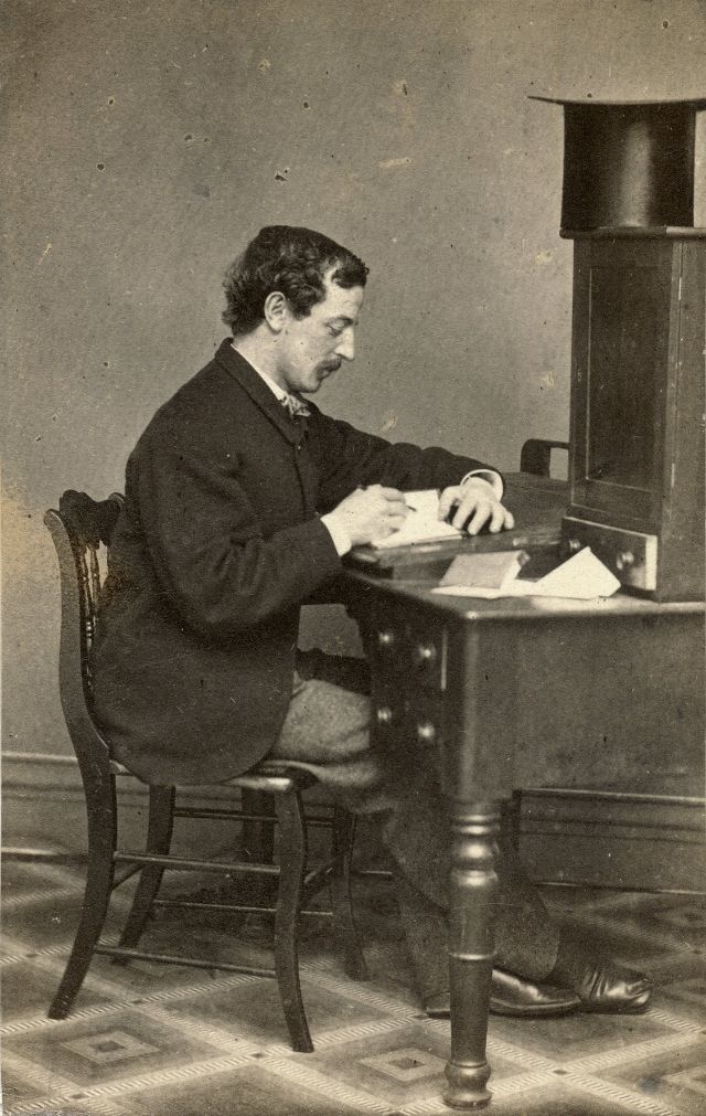 #7 A man sits at a desk and writes in a small volume similar in size and thickness to a journal. His top hat rests on top of the cabinet mounted to the desktop