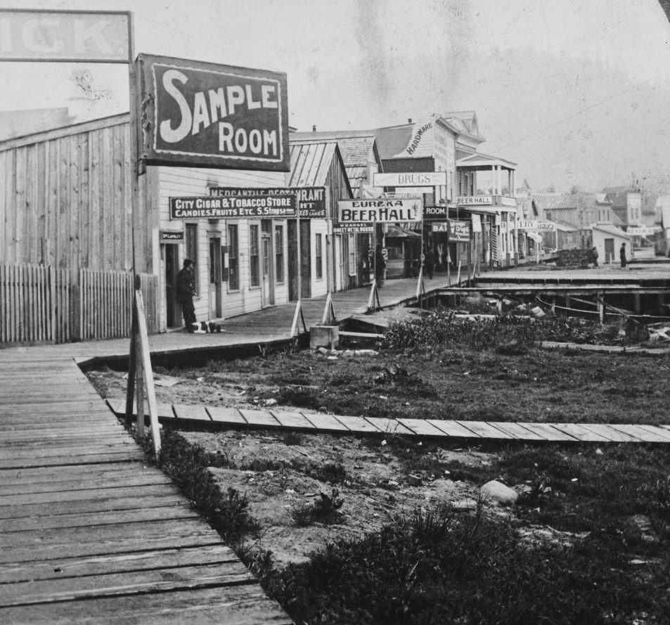 #50 Main Street in Wrangell, Alaska, featuring saloons and totem poles, during the Klondike Gold Rush, circa 1900.