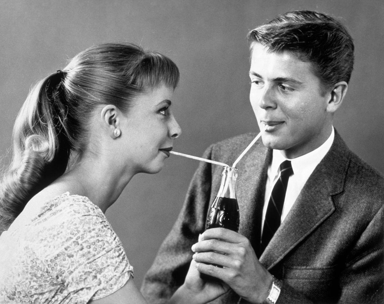 #64 Teenage couple sharing a single bottle of soda with two straws, 1950s.