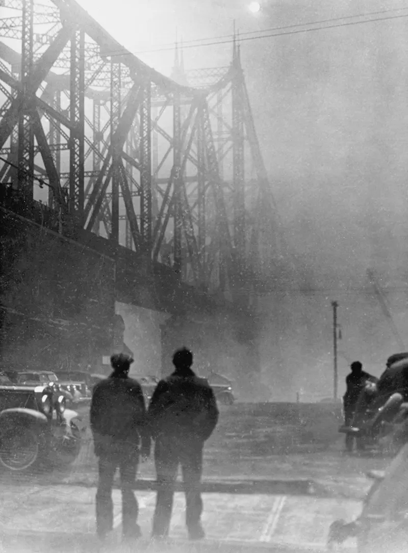 #6 Two men by traffic under a bridge, third man leaning over a car, sun visible through smoke.