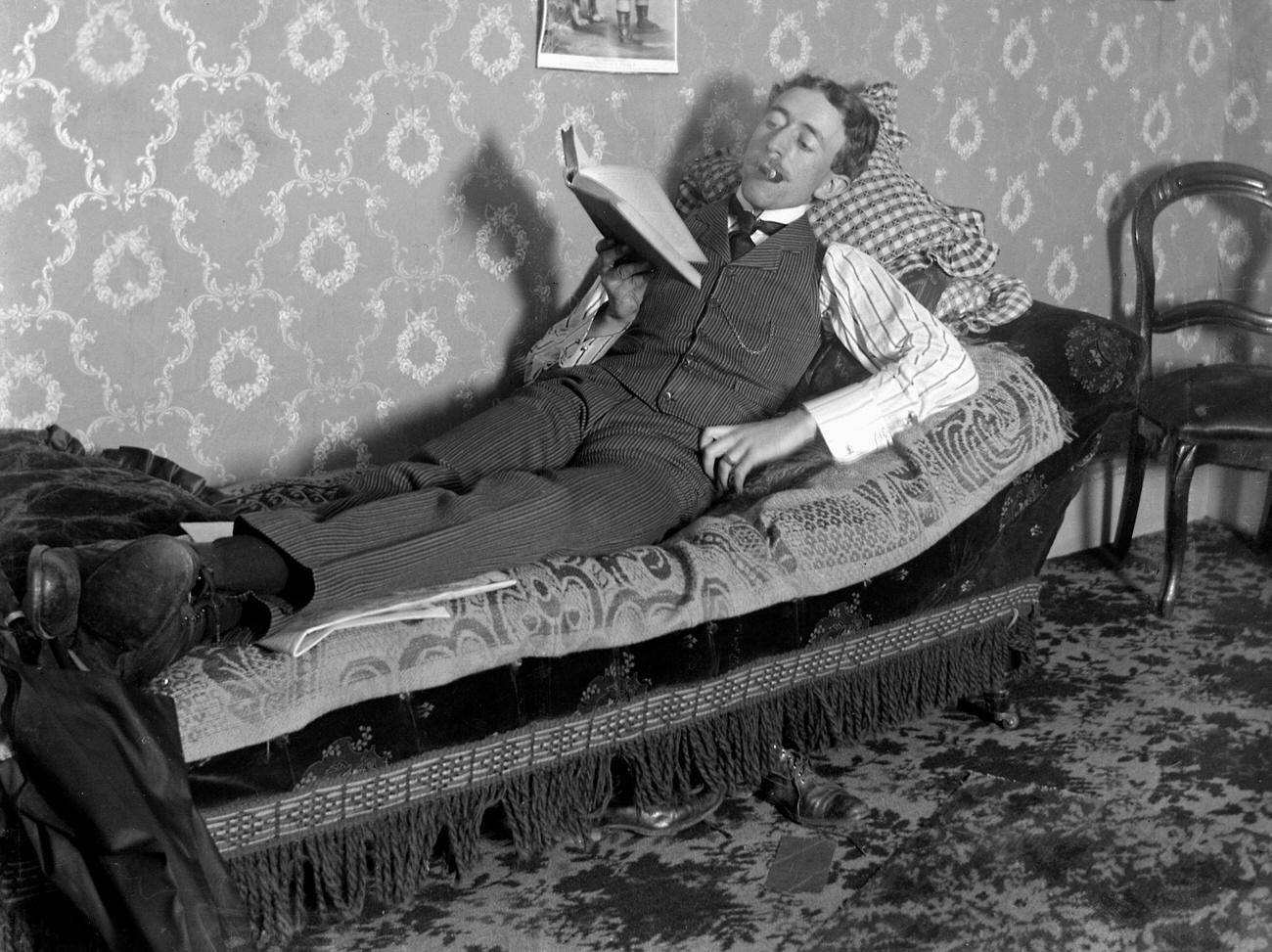 #72 A man reading and smoking on a fainting couch, circa 1900.