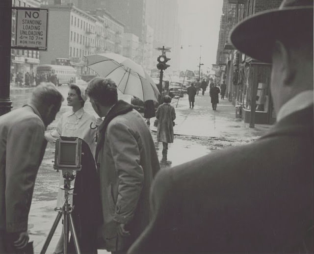 #13 Woman being photographed in the rain on a street corner, 1954