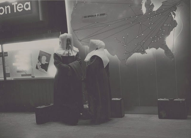 #22 View of two nuns next to map of the United States with route markers and lights, suitcases on ground, November 1957