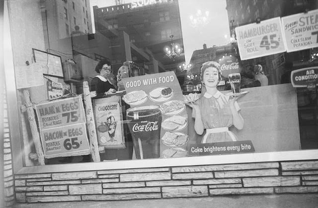 #34 Woman at cash register viewed through diner show window with buildings reflected, February 1959