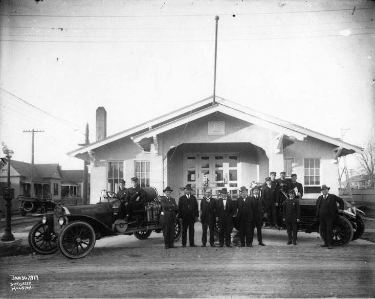 #62 Men between two Houston firetrucks with steering wheels on the right, Fire Station No. 12 in the background, 1917.