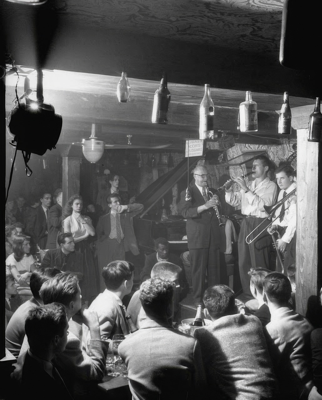 Jam session in shadowy cellar of Vieux Colombier attracts a crowd of Americans (foreground). Their compatriot, Clarinetist “Mezz” Mezzrow (left), is the big attraction. On such forays, the boys save money by dividing a bottle of champagne eight ways. In Paris, American teen-agers ignore Eckstine and Sinatra records as démodé, prefer French Crooners Charles Trenet and Yves Montand. They have a favorite French tune, Fou de Vous (Crazy About You).