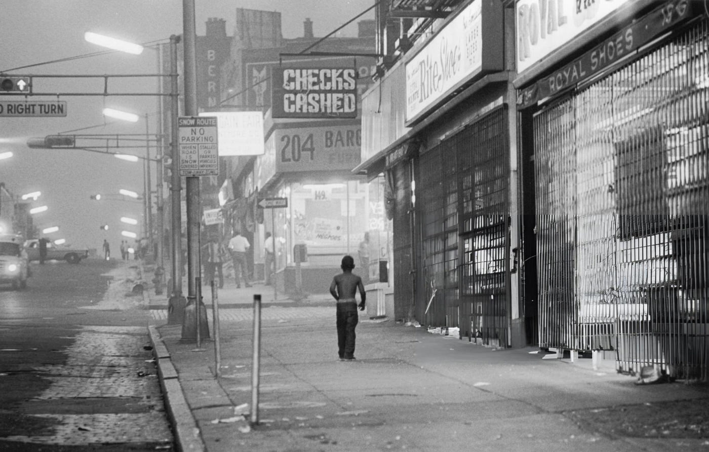 #13 A young boy walks down the sidewalk past shuttered storefronts towards a furniture store