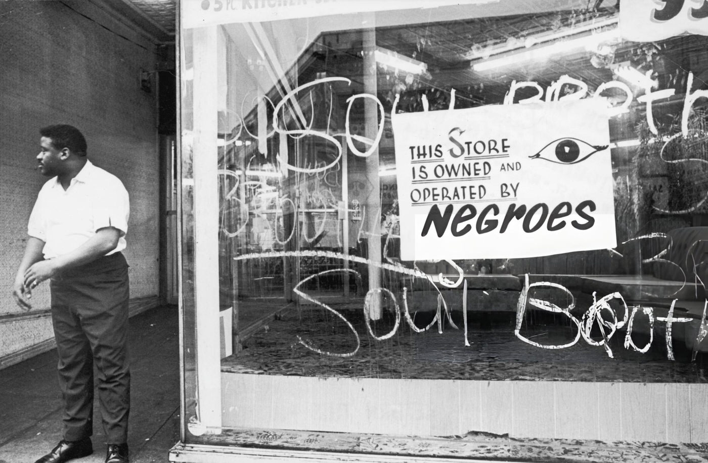 #24 An African-American storekeeper stands outside his shop, which has a sign reading, ‘This store is owned and operated by Negroes.