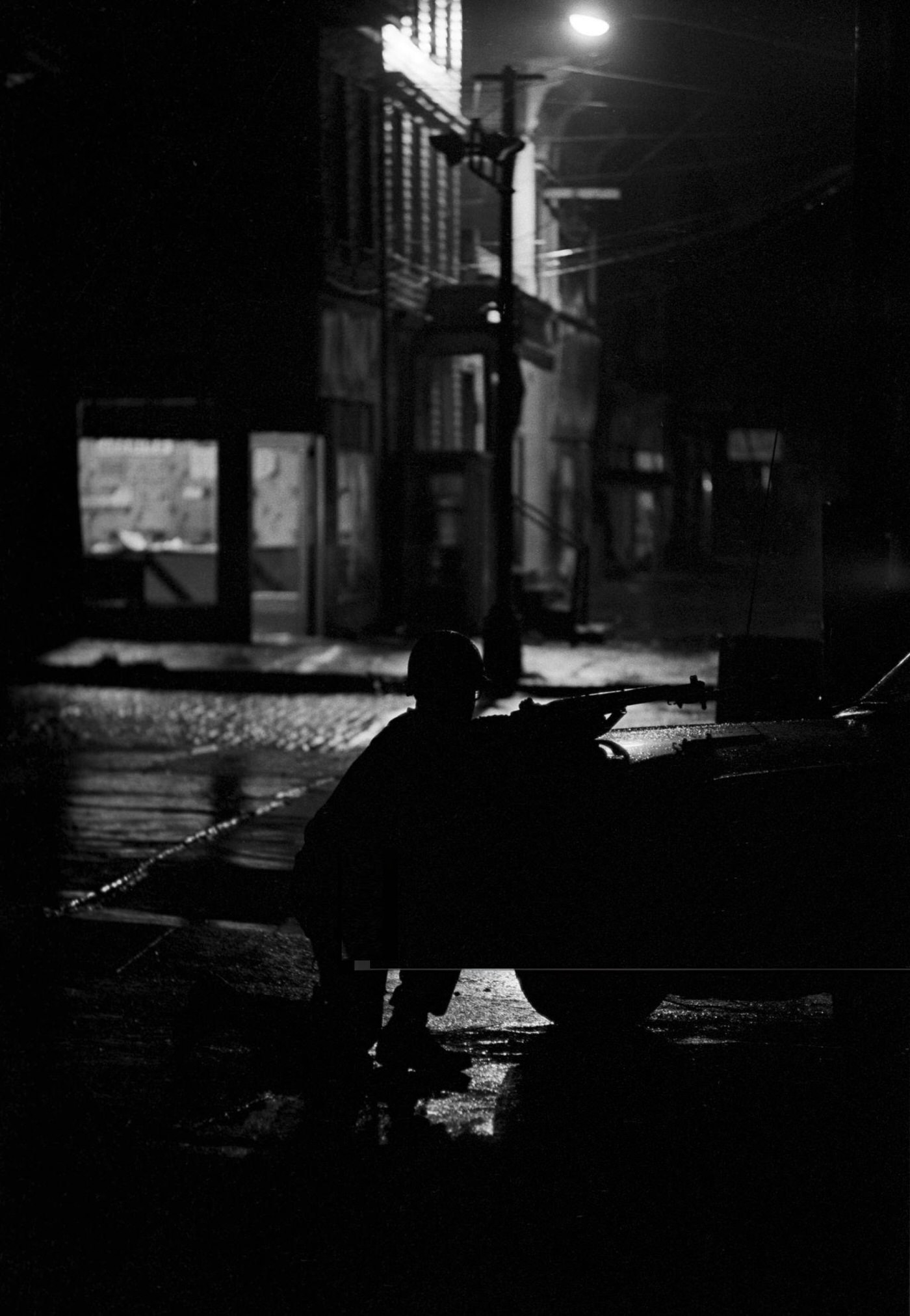 #50 A National Guard soldier crouched behind a car, gun pointed towards the street at night