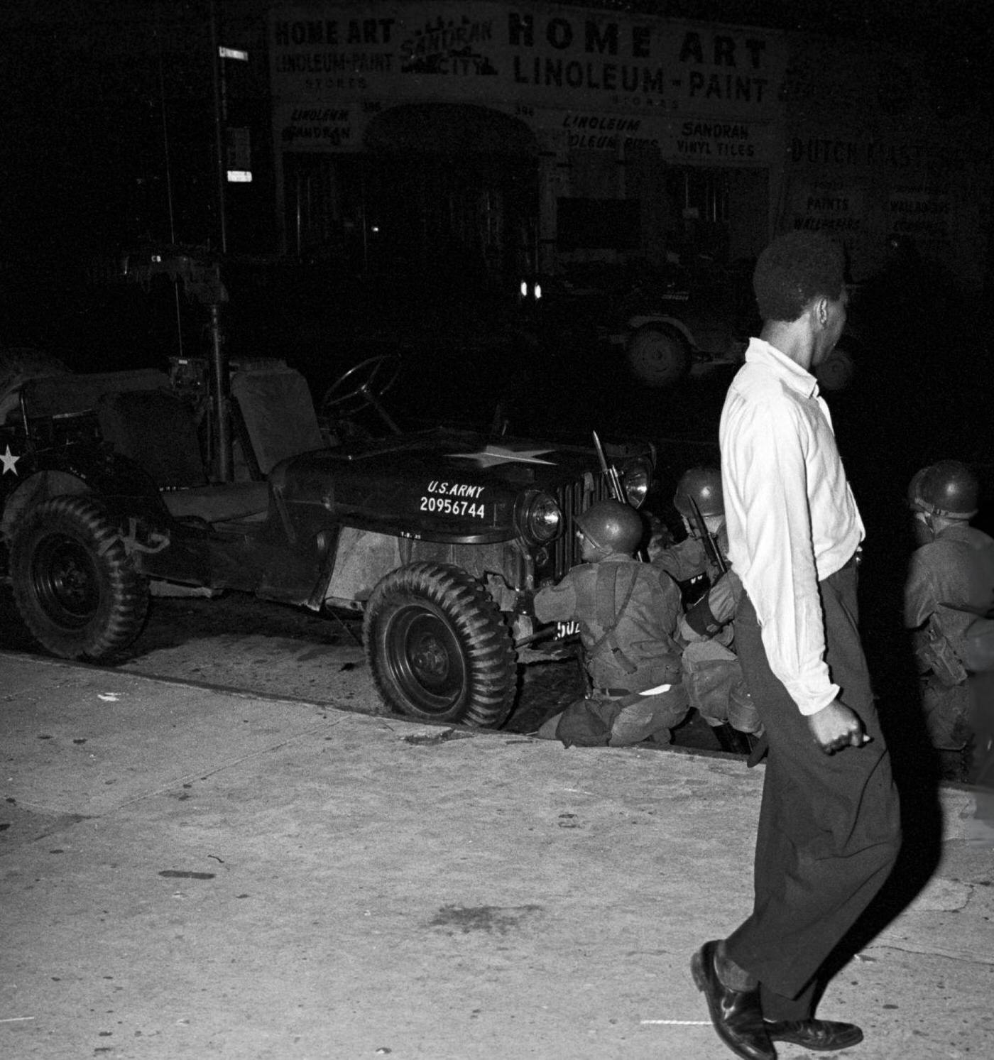 #51 A young African American man walking on a sidewalk looks at a group of National Guard soldiers, 1960s.