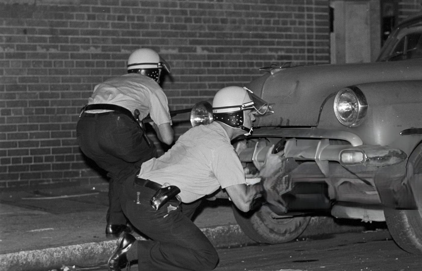 #58 Two police officers in riot helmets take cover behind a parked car