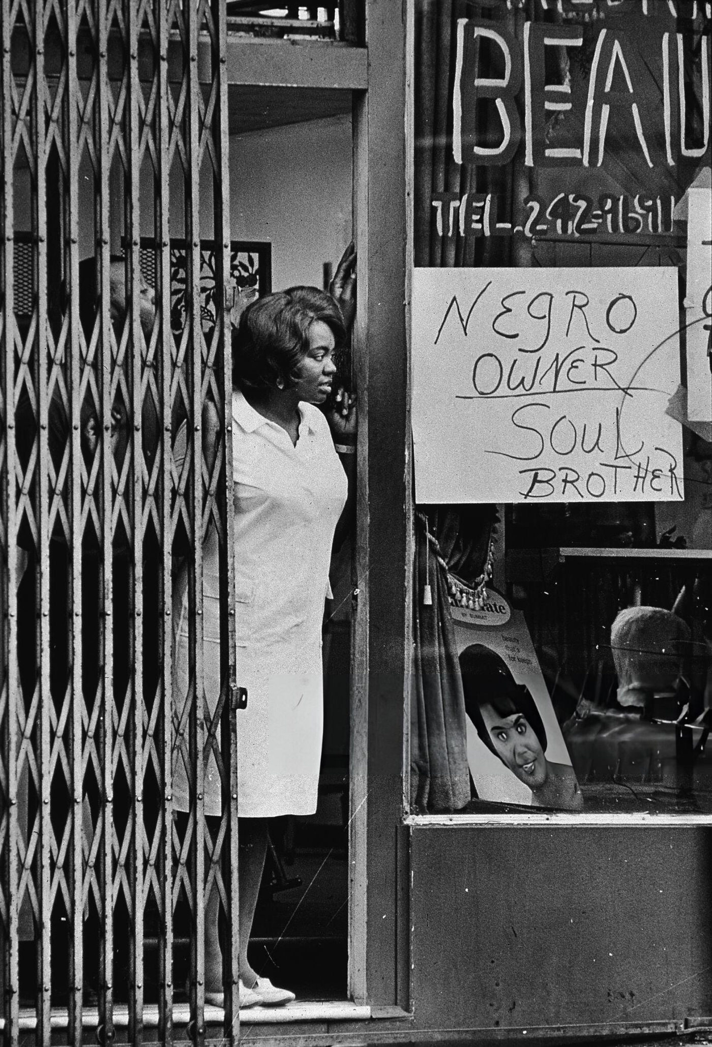 #5 A Black woman peers from beside a gate in the door of a store with a sign in the window reading ‘Negro owner,’ 1967