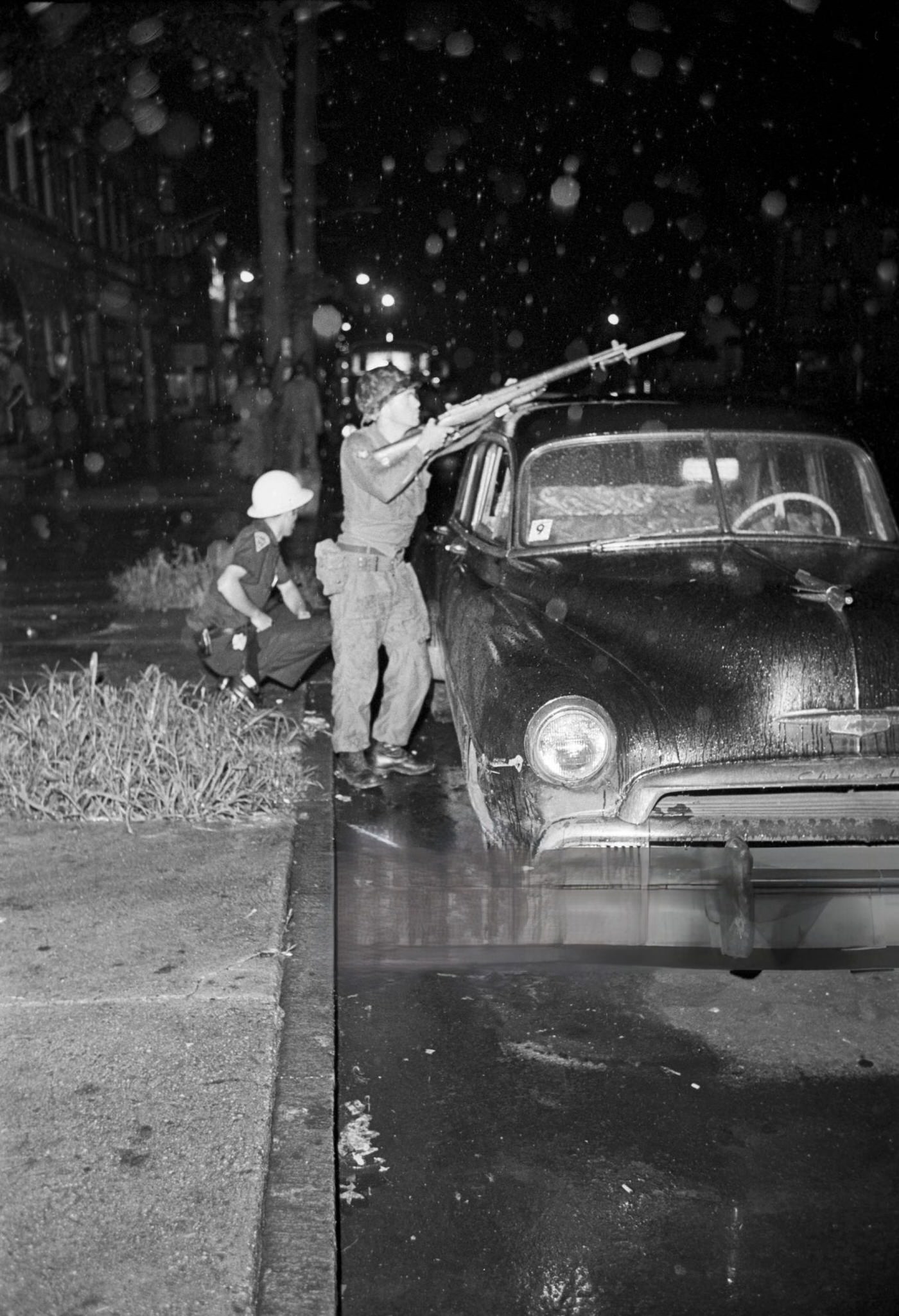 #64 A National Guard soldier stands with a gun pointed towards the street next to a police officer crouched behind a jeep