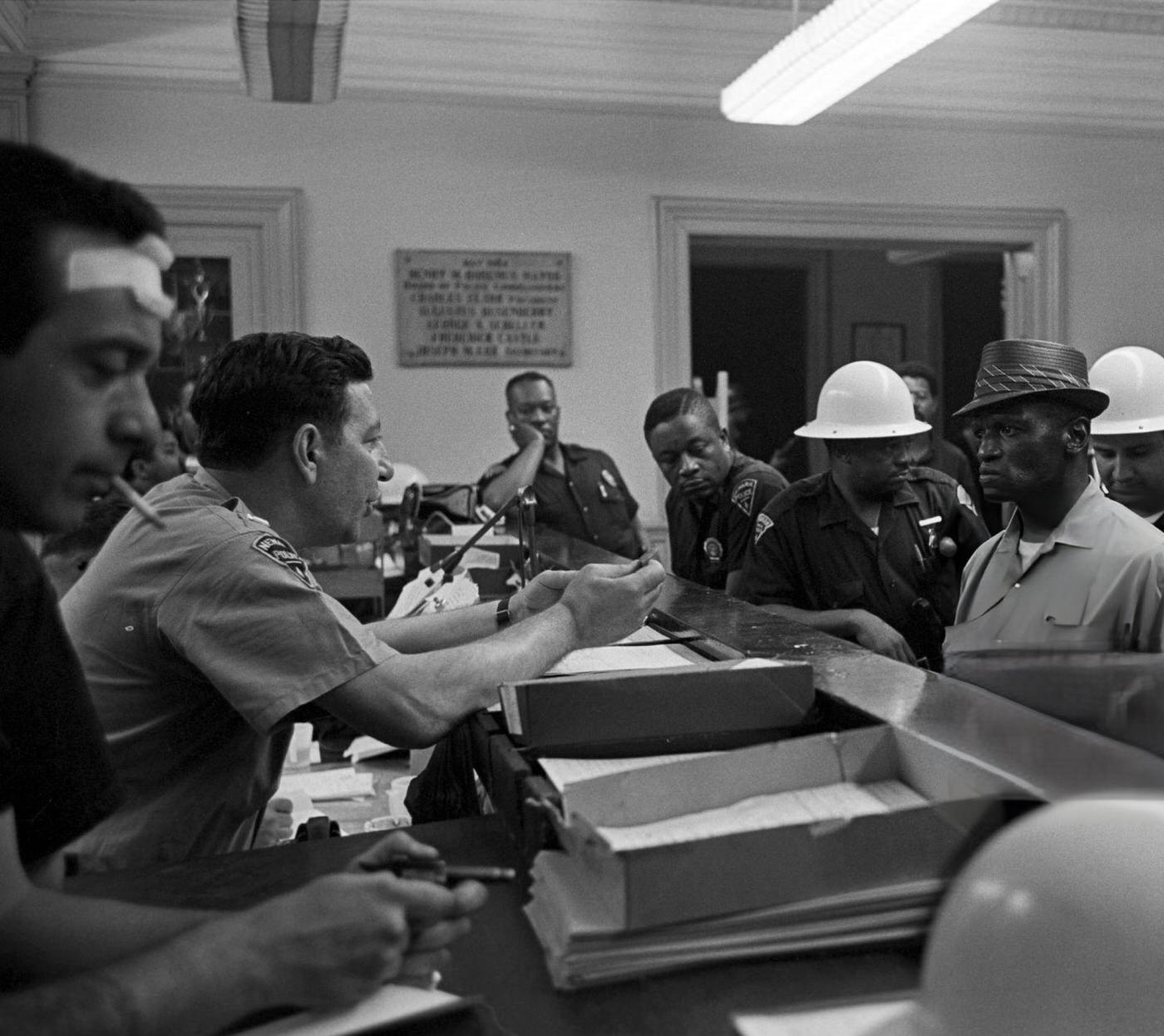 #65 An African-American man in a hat under arrest, standing in front of a counter surrounded by Newark police officers, 1960s.