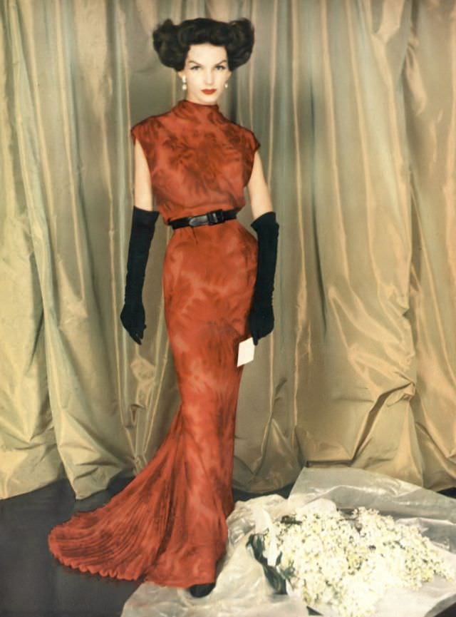 #19 Joanna McCormick in a garnet red print chiffon sheath, bloused over a leather belt, an air-vent in the back and a fan pleat train, by Balmain, 1957.