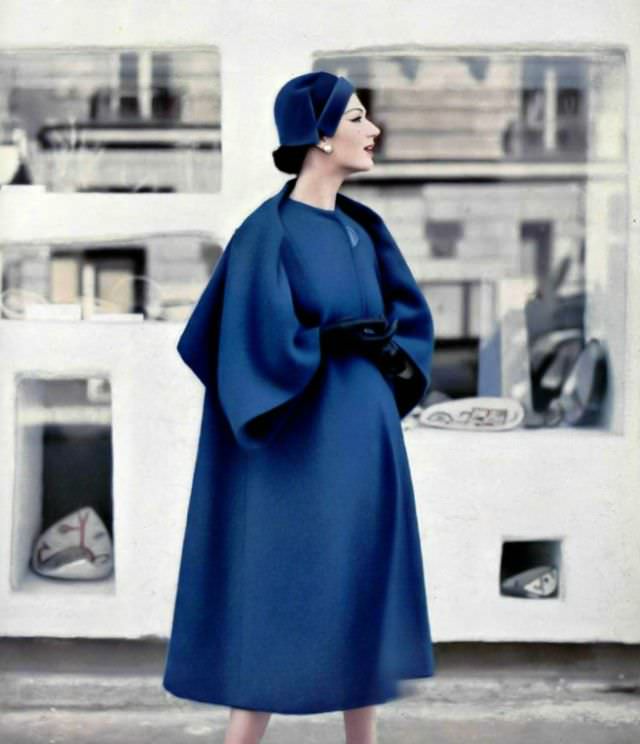 #26 Simone d’Aillencourt in a roomy blue coat whose effect of a short cape at the back is given by the cut and sleeve setting, collarless and one large button-closure, by Pierre Balmain, 1957.