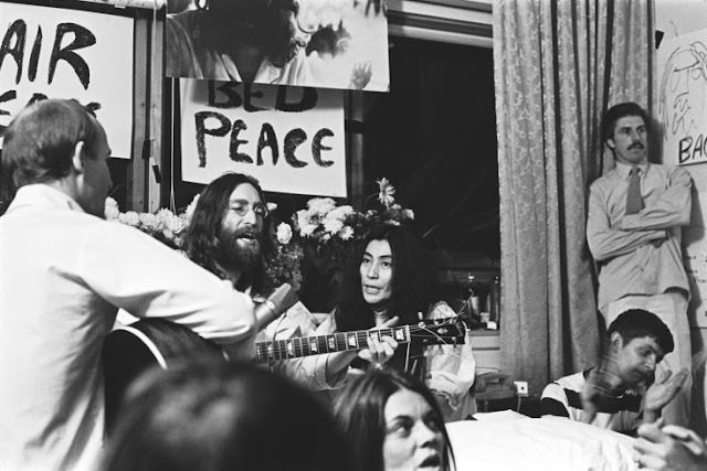The couple sits among friends Tommy Smothers, far left, and Derek Taylor, standing far right, singing “Give Peace a Chance.”