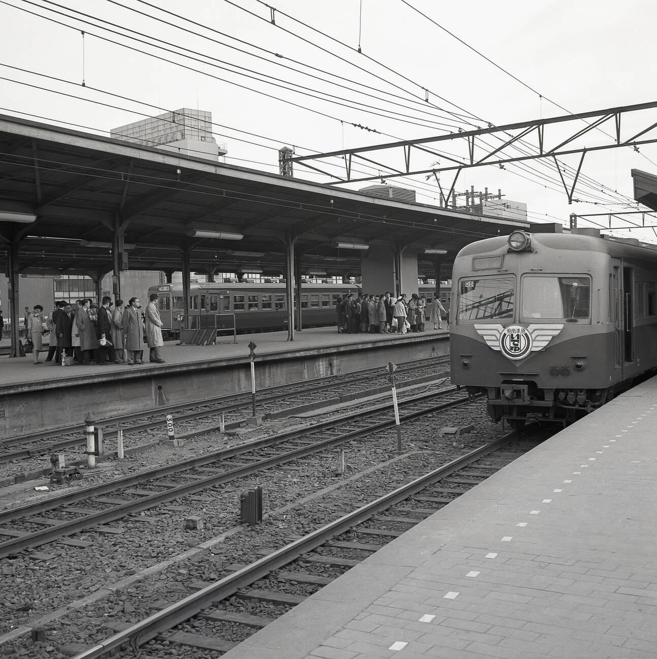 #58 An overground train arrives on one platform, while Japanese rail commuters wait for their train to arrive, 1950s.