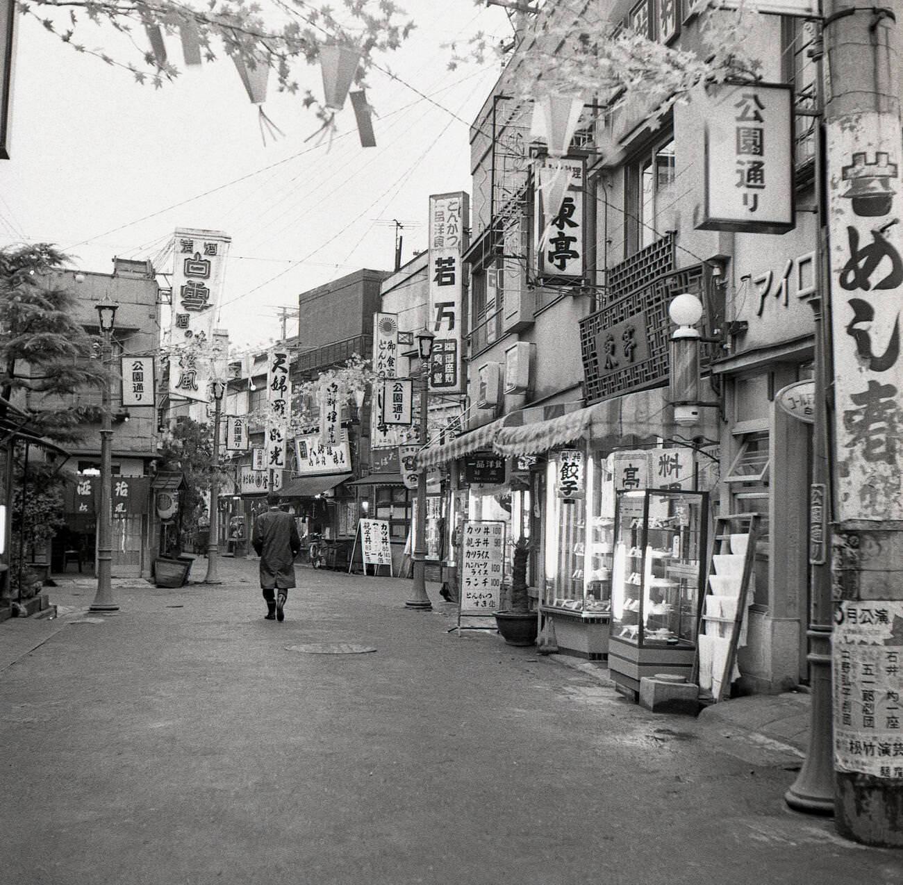 #60 A man walking down pass the retail stores and buildings in a quiet side-street in Tokyo, 1950s.
