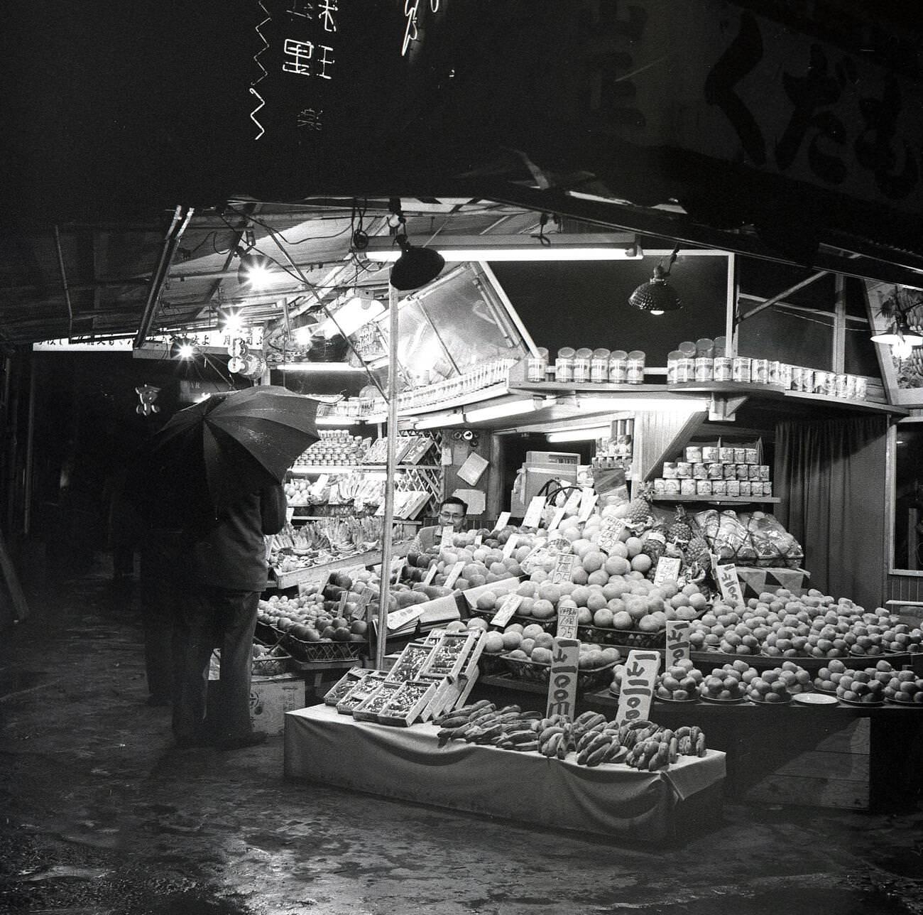 #78 A wet evening and a man with an umbrella at an outdoor street stall selling fruit in Tokyo, 1950s.