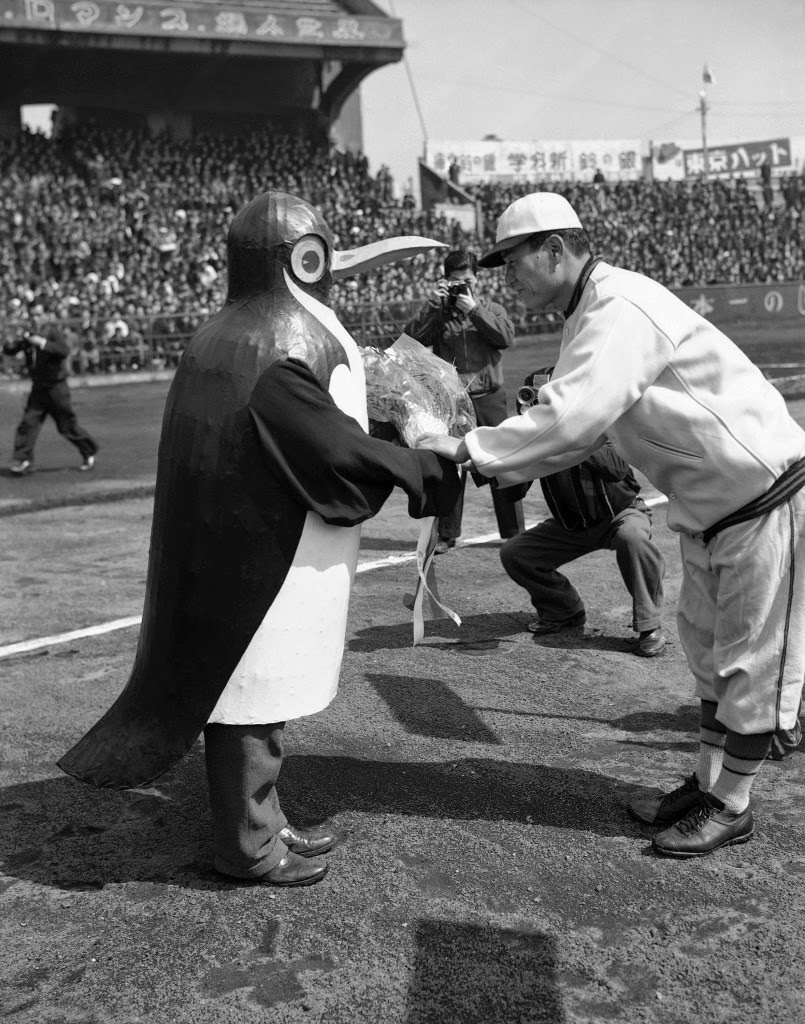 #82 As the baseball season opened in Japan, a penguin-costumed attendant presents flowers to the manager of one of the eight teams in the league in Tokyo, 1949.