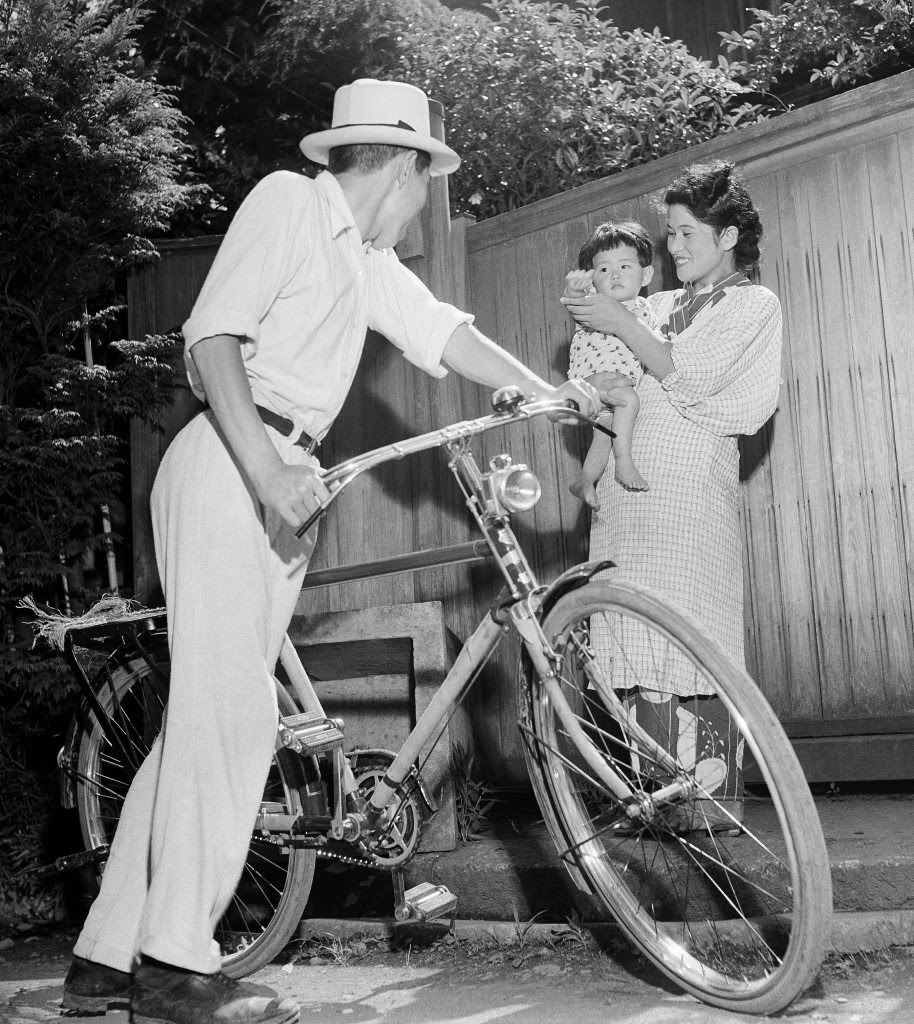 #88 Baby Misuko, held by her mother Umeko, waves goodbye to papa-san as he goes to work on his bicycle, from their home in Tokyo, 1948.