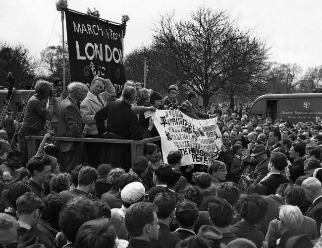 #24 Two survivors of the Hiroshima atomic bomb hold up a banner as they join leaders of the Easter Ban-the-Bomb march on the dais in London’s Hyde Park at the mass rally climaxing the 50-mile march from Aldermaston, 1962.
