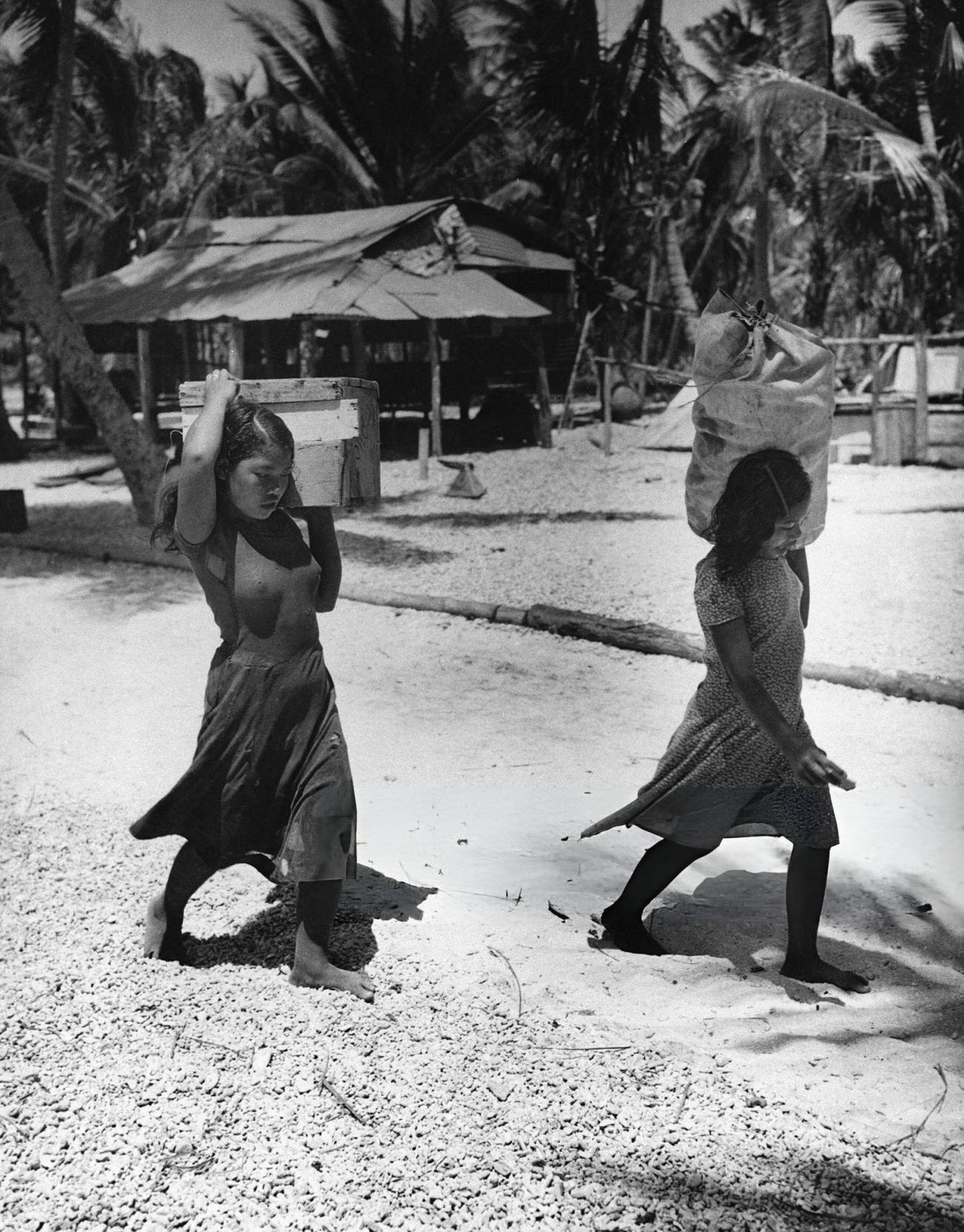 #20 Two Bikini Atoll girls trudge away from their village carrying their possessions on their shoulders, 1946.