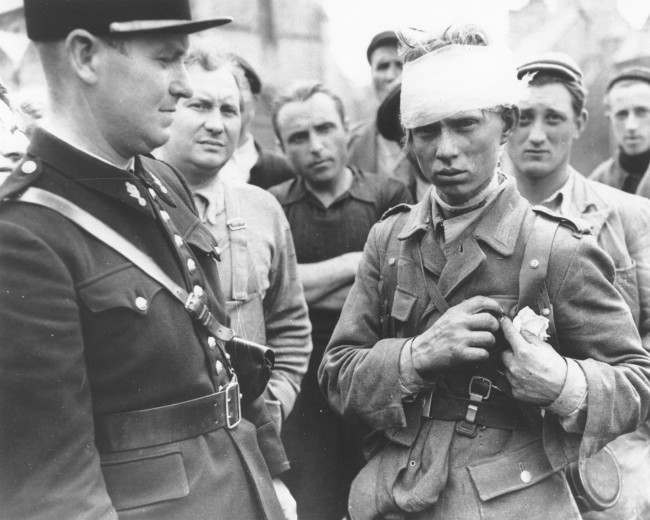 #21 A wounded German soldier who surrendered to the Allied invasion forces, stands surrounded by a crowd of civilians, 1944.