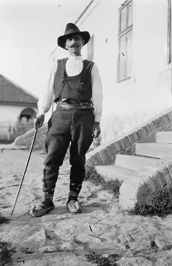#27 Serbian man wearing hat, vest, belted pants, and pointed toed shoes with cane standing outside of building in Yugoslavia, 1880-1924.