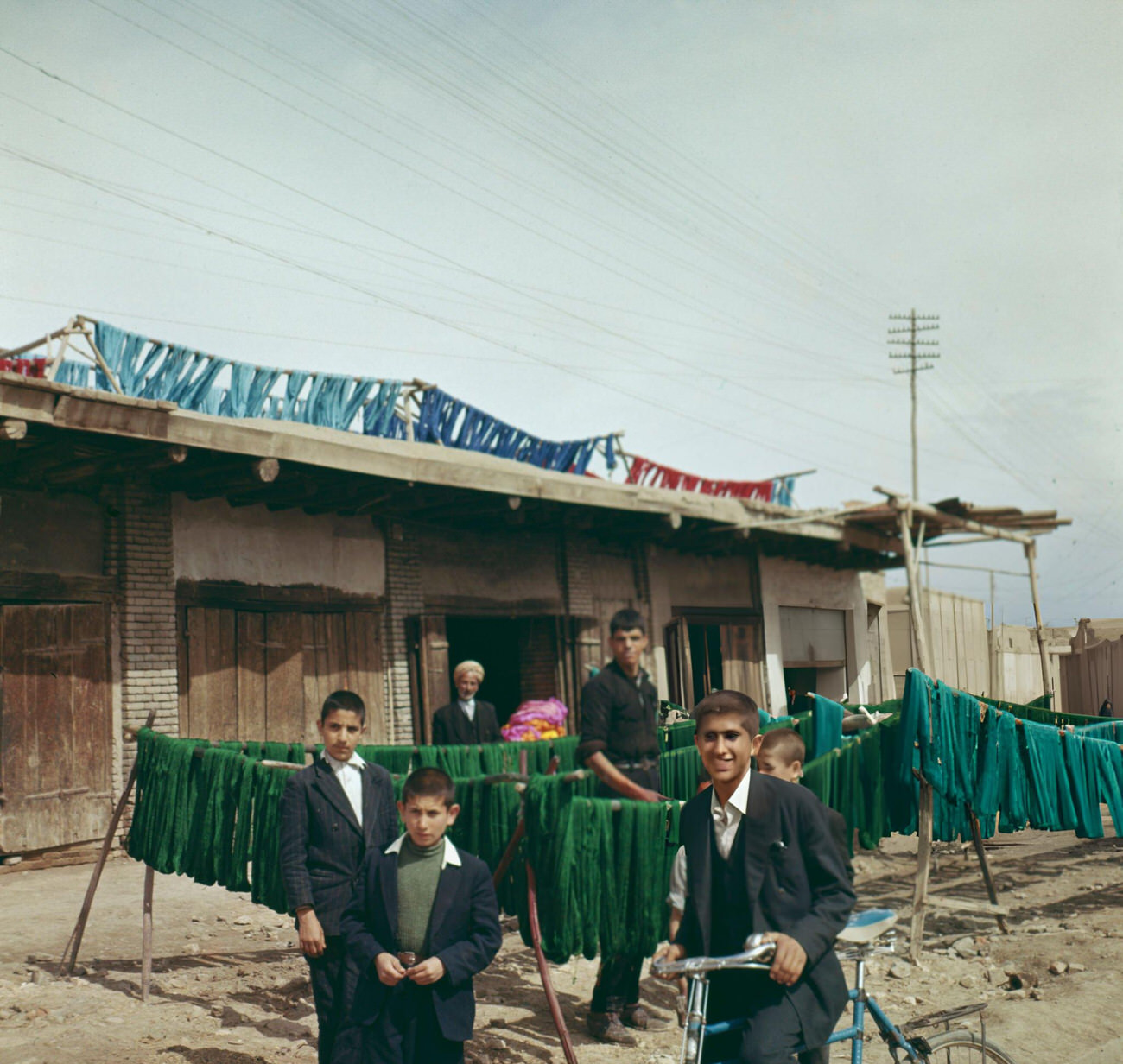 #28 Local boys congregate in front of carpet wools being hung out to dry after dyeing in various colors at a workshop on a street in Iran, 1963.