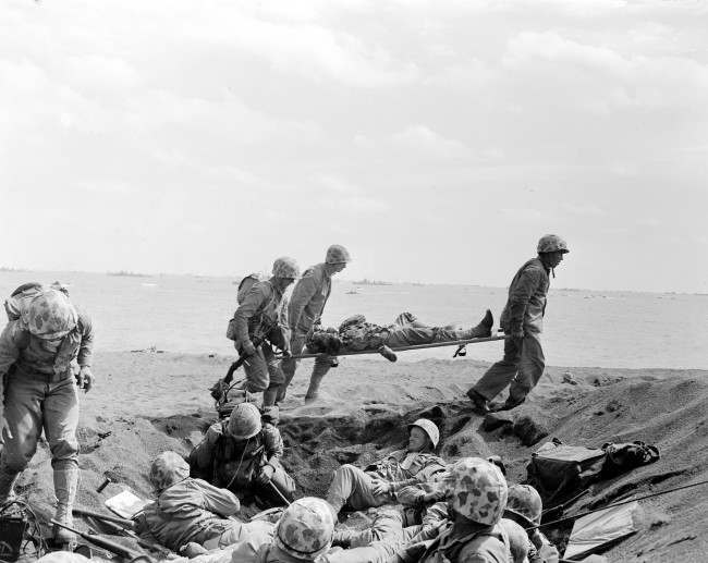 #23 Corpsmen carry a wounded Marine on a stretcher to an evacuation boat on the beach at Iwo Jima while other Marines huddle in a foxhole during the invasion of the Japanese Volcano Island stronghold, 1945. A search team is on the island looking for a cave where the Marine combat photographer who filmed the famous World War II flag raising is believed to have been killed in battle nine days later, military officials said Friday, June 22, 2007.